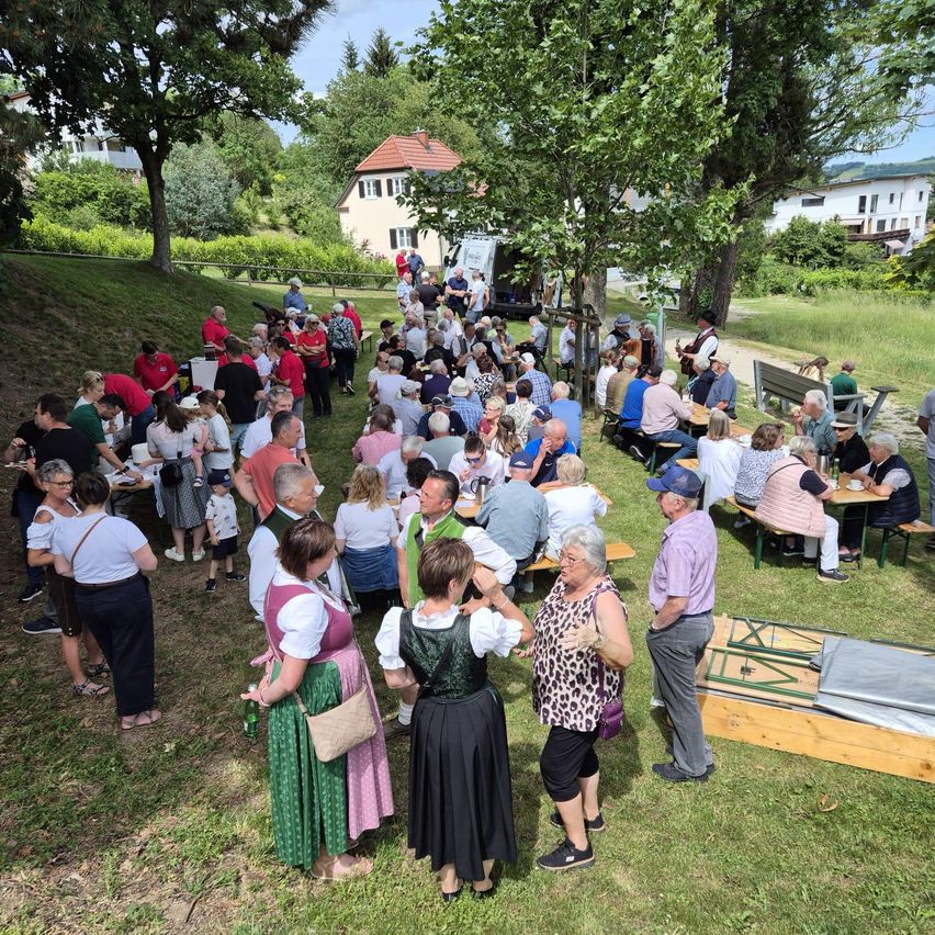 Eine große Gruppe von Menschen in traditioneller Kleidung versammelt sich auf einem grasigen Hang, einige sitzen an Tischen, andere stehen und unterhalten sich. Bäume bieten Schatten, und ein Gebäude ist im Hintergrund zu sehen.