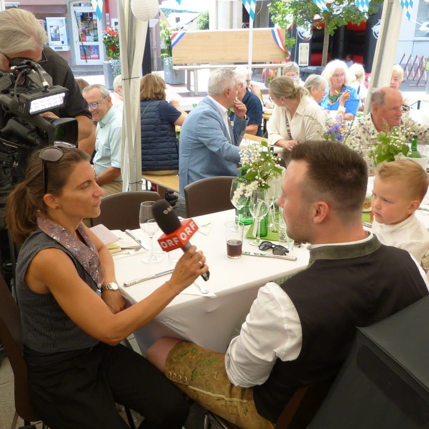 Eine Frau hält ein Mikrofon und interviewt einen Mann an einem Tisch in einem Restaurant. Hinter ihnen sitzen mehrere Menschen an Tischen und essen. Ein Kameramann hält die Szene fest.
