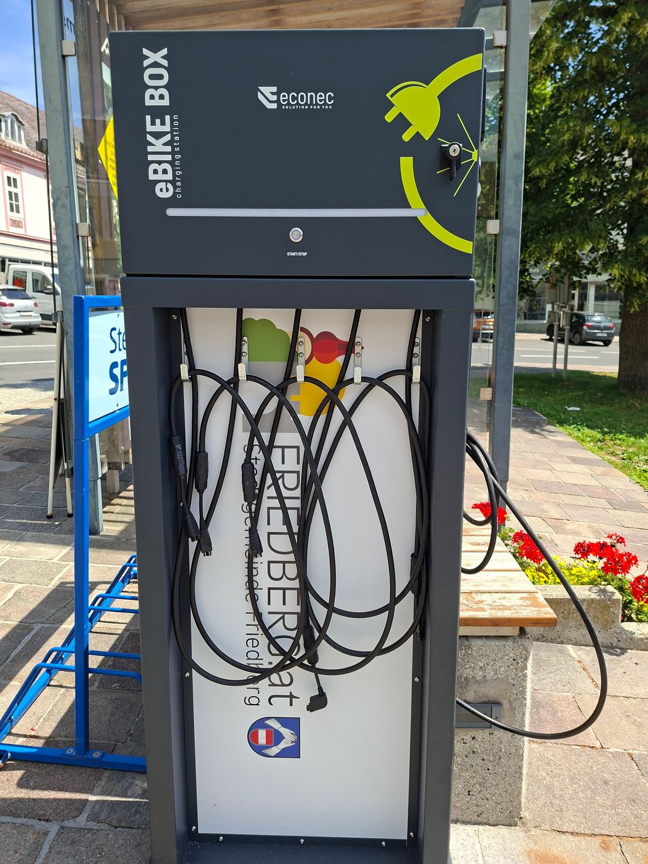 A charging station for electric bikes is outdoors. The station is gray with a yellow logo. Several charging cables hang from it.