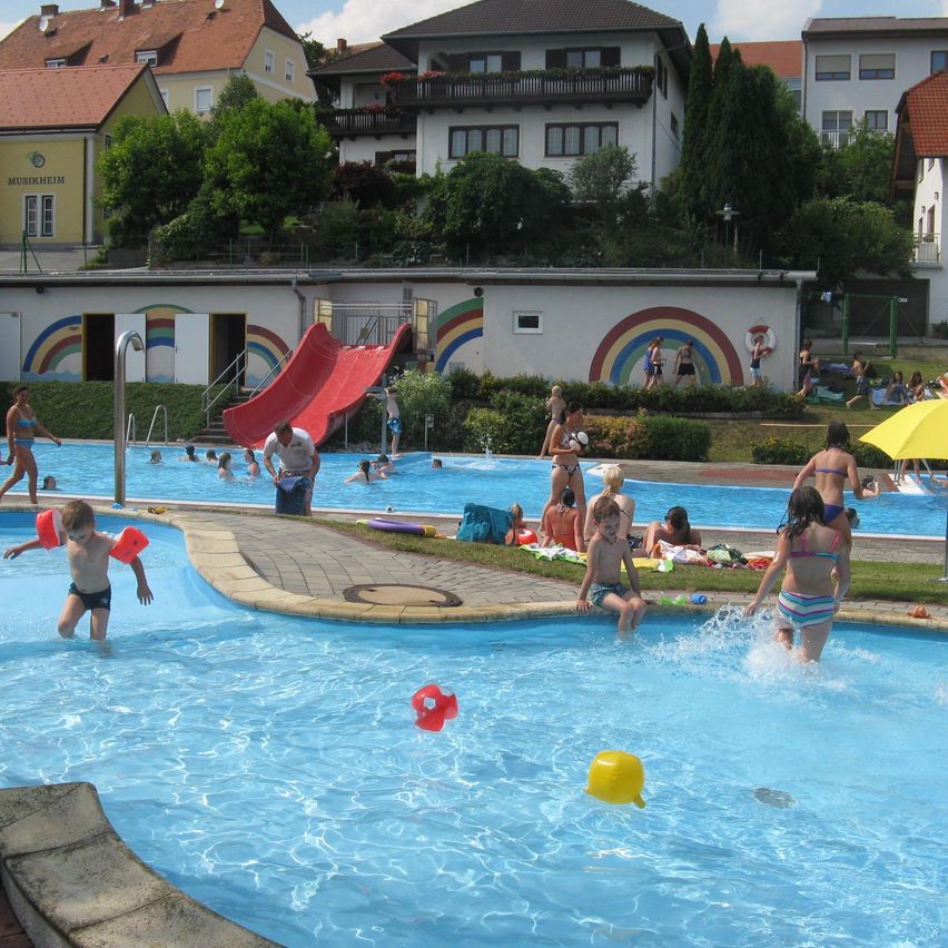 A group of children playing in a swimming pool. A red slide and rainbow decorations are visible.