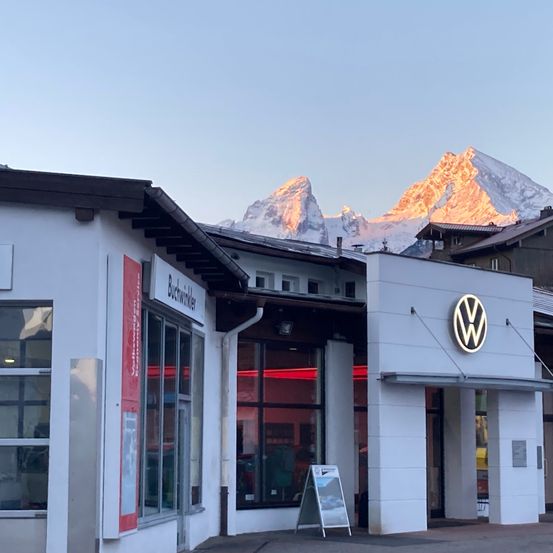 Ein Volkswagen-Händler steht in einer Bergregion mit schneebedeckten Bergen im Hintergrund. Das Gebäude hat ein modernes Design mit Glasfenstern und einem Logo über dem Eingang.