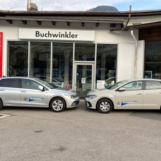 Zwei silberne Volkswagen-Autos sind vor einem Gebäude namens Buchwinkler geparkt und zeigen ihr Design und ihre Marke. Die Umgebung umfasst einen klaren Himmel und entfernte Berge.