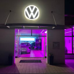 Der Eingang zu einem Volkswagen-Händler ist nachts beleuchtet. Das Logo ist deutlich über dem Eingang zu sehen. Ein Auto ist drinnen geparkt.