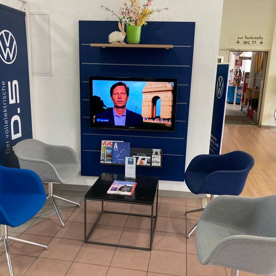 Ein Wartebereich mit Fernseher, Zeitschriften und einem Tisch. Es gibt blaue und graue Stühle, eine blaue Wand und ein VW-Schild.