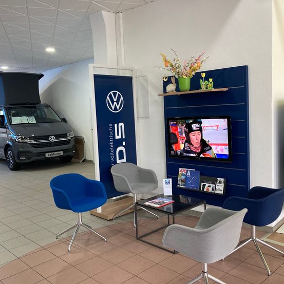 Ein Wartebereich in einem Autohaus zeigt einen Volkswagen-Transporter, Stühle, einen Couchtisch und einen Fernseher. Es gibt ein VW-Banner, ein Regal mit einer Topfpflanze und einen Fernseher, der ein Rennen zeigt.