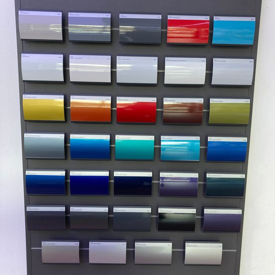 Eine Sammlung verschiedener Farbmuster wird auf einem grauen Brett angezeigt. Jedes Muster hat ein Etikett mit einem Farbnamen und -code und zeigt eine Reihe von Farben von hellgrau bis dunkelviolett.