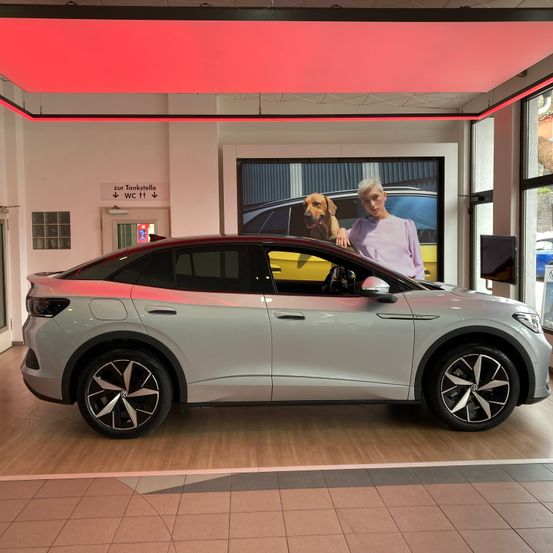 Ein graues Auto wird in einer Ausstellung gezeigt. Eine Frau mit einem Hund steht hinter dem Auto. Eine rote Leuchte ist an der Decke und ein Poster ist hinter dem Auto.