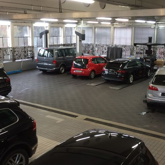 Ein überdachter Parkplatz mit mehreren geparkten Autos, darunter ein rotes Auto, ein grauer Van und ein schwarzes Auto auf einem Lift. Der Bereich hat große Fenster, Deckenleuchten und geflieste Böden.