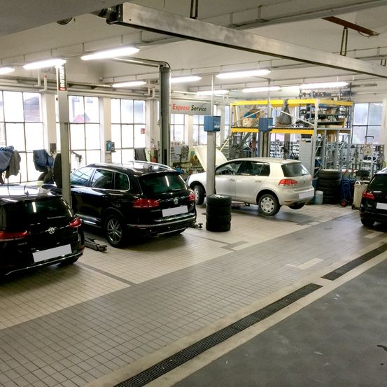 Eine Autowerkstatt mit mehreren geparkten Autos zur Wartung. Ein weißes Auto mit offenem Motorraum und ein schwarzes Auto mit angehobener Rückseite. Reifenstapel und Regale mit Gegenständen im Hintergrund.
