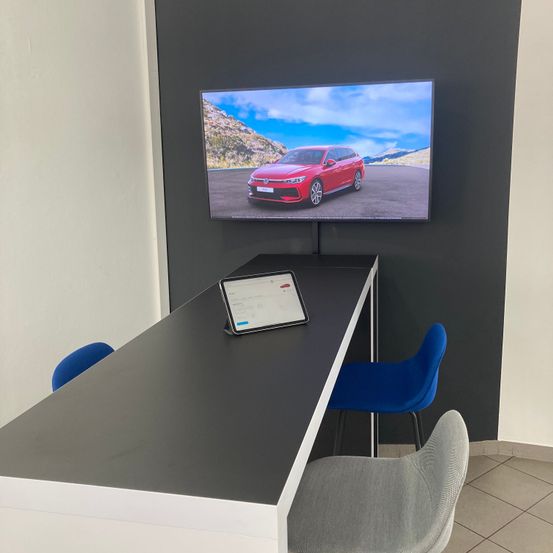 Ein rotes Auto wird auf einem montierten Bildschirm in einem Besprechungsraum mit einem schwarzen Tisch, zwei blauen Stühlen und einem Tablet angezeigt.