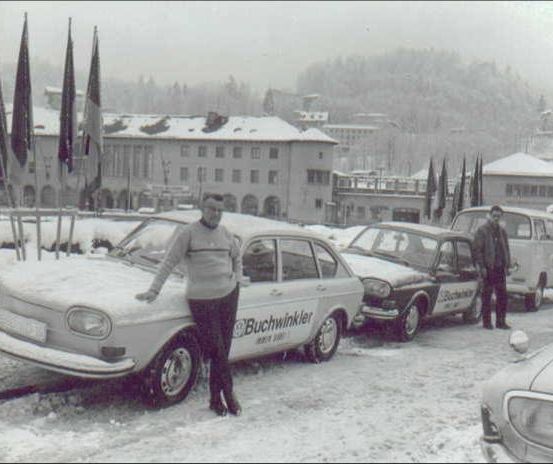 Ein Schwarz-Weiß-Foto zeigt eine verschneite Straße mit mehreren geparkten Autos. Ein Mann lehnt sich an ein weißes Auto, während ein anderer Mann neben einem Van steht. Im Hintergrund sind Fahnen und Gebäude zu sehen.