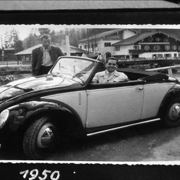 Ein Schwarz-Weiß-Foto von 1950 zeigt einen Mann in einem Cabrio, neben dem ein anderer Mann steht. Das Cabrio ist vor einem Gebäude geparkt.