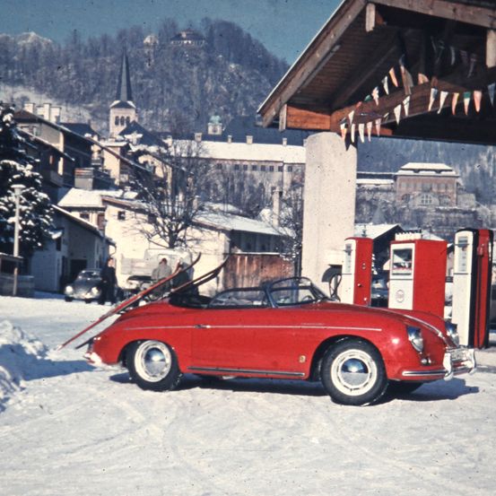 Ein roter Cabrio-Sportwagen mit Skiern auf dem Dach steht vor einer Tankstelle in einer verschneiten Stadt.