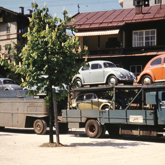 Ein Lkw mit mehreren Oldtimern steht vor einem Gebäude mit rotem Dach. Die Autos sind silbern, gelb und orange. Ein kleiner Baum steht vor dem Lkw.