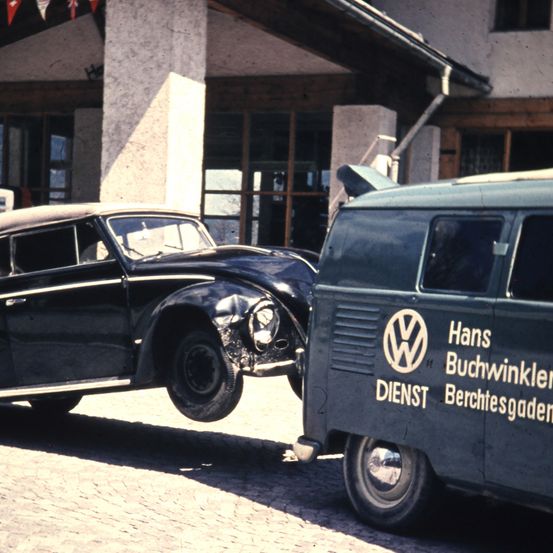 Ein altes schwarzes Oldtimer-Auto ist vor einem Gebäude geparkt. Es scheint in einen Unfall verwickelt gewesen zu sein. Daneben steht ein Volkswagen-Bus mit den Worten 'Hans Buchwinkler Dienst Berchtesgaden'.