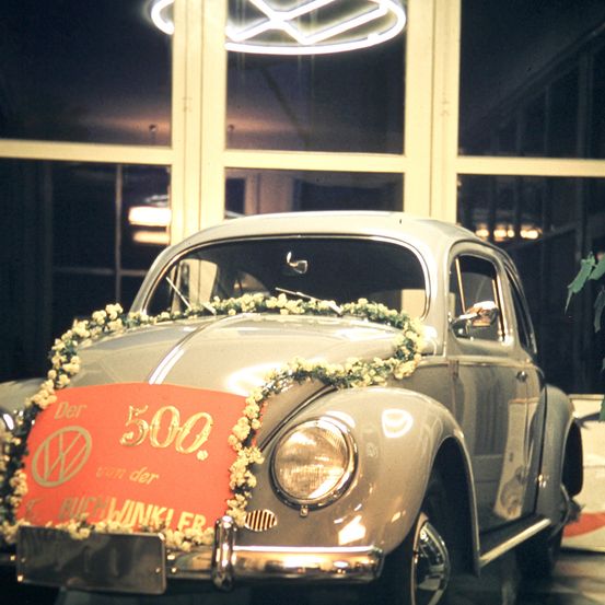 Ein Vintage grauer Volkswagen Käfer mit einem roten Banner, das 'Der 500' sagt, ist in einer Ausstellung geparkt. Das Auto ist mit weißen Blumen geschmückt.