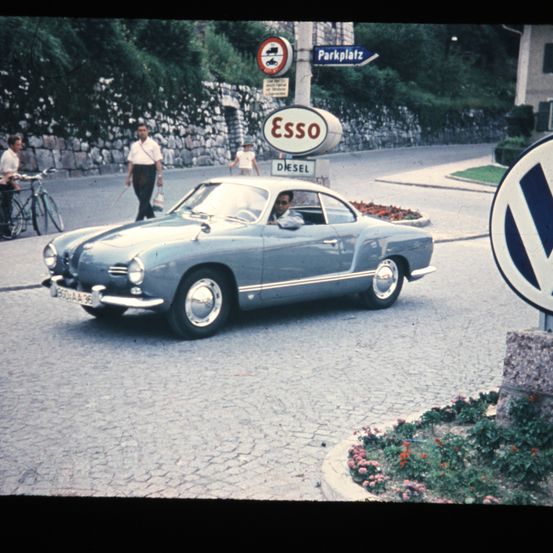 Ein klassischer VW Karmann Ghia fährt eine Straße hinunter, in der Nähe eines Esso-Diesel-Schildes. In der Nähe gehen Menschen und fahren mit dem Fahrrad.