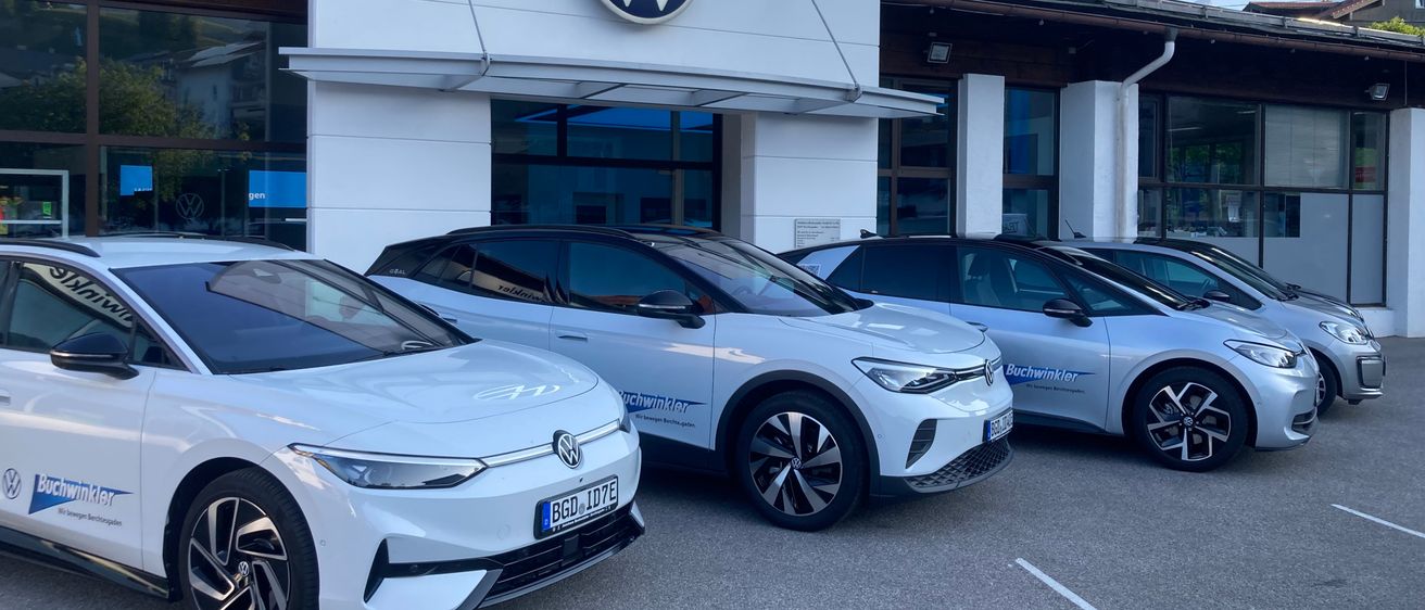 Ein weißes Volkswagen-Autohaus ist zu sehen, vor dem mehrere Elektroautos geparkt sind. Das Autohaus hat ein großes VW-Logo über dem Eingang. Die Autos sind in einem Parkplatz mit weißen Markierungen aufgereiht.
