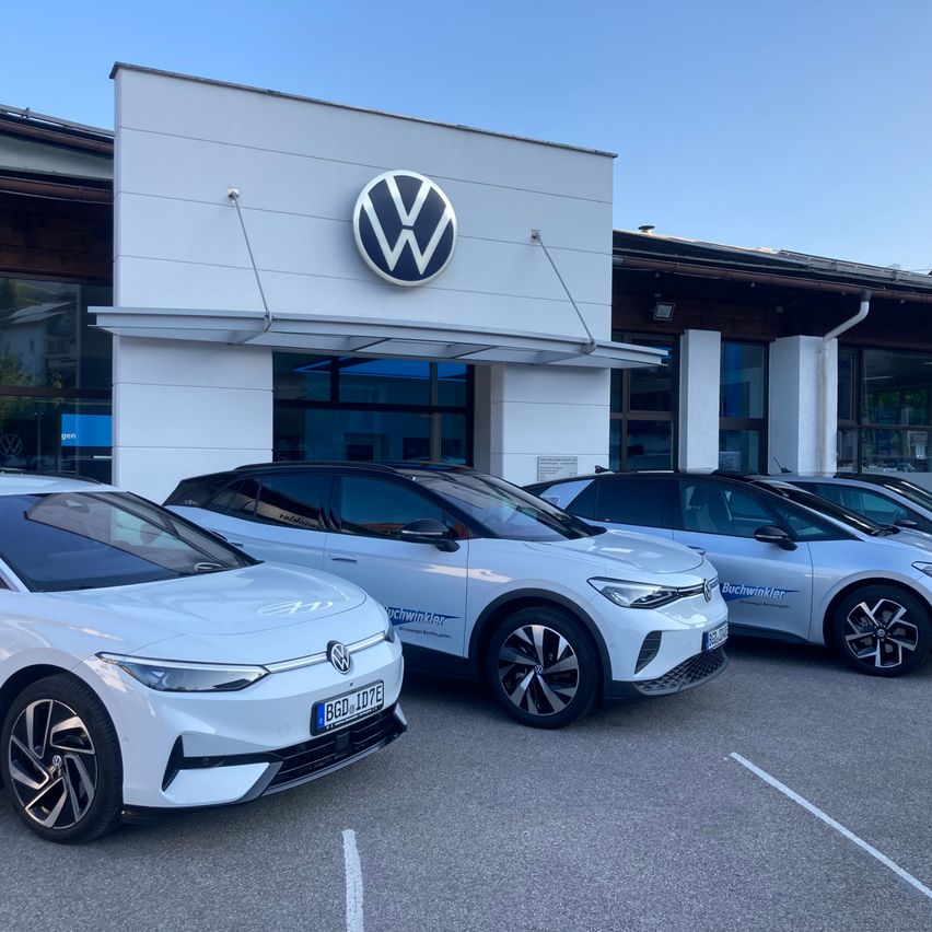 Ein weißes Volkswagen-Autohaus ist zu sehen, vor dem mehrere Elektroautos geparkt sind. Das Autohaus hat ein großes VW-Logo über dem Eingang. Die Autos sind in einem Parkplatz mit weißen Markierungen aufgereiht.