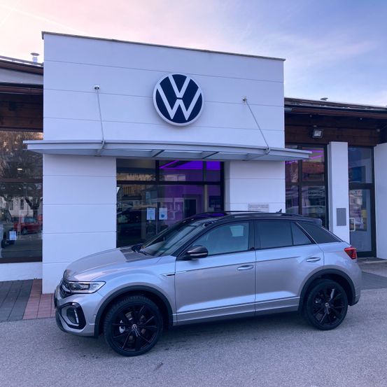 Ein silberner Volkswagen SUV steht vor einem modernen weißen Händler mit einem Volkswagen-Logo über dem Eingang.
