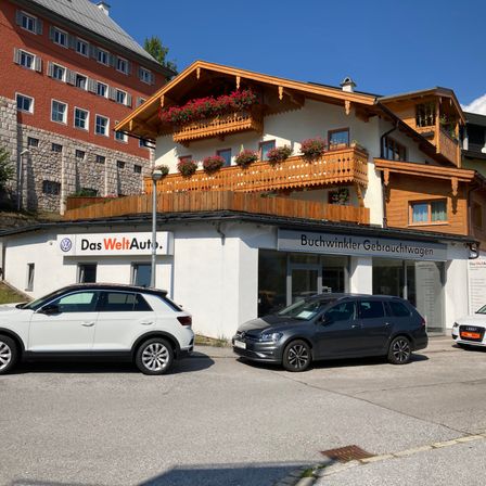 Zwei Autos sind vor einem Gebäude geparkt. Das Gebäude hat zwei Schilder, eines davon steht für Das WeltAuto und das andere für Buchwinkler Gebrau. Hinter dem Gebäude befindet sich ein Haus mit Balkonen, die mit roten Blumen geschmückt sind.