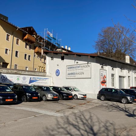 Ein Gebäude mit einem Schild, das Gebrauchtwagen Jahreswagen Volkswagen anzeigt, mit mehreren Autos davor geparkt. Ein Gebäude im Bau dahinter und Bäume im Hintergrund.