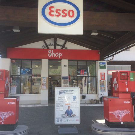 Eine Tankstelle mit einem ESSO-Schild über dem Laden. Vorne stehen zwei ESSO-Kraftstoffspender. Ein Schild steht zwischen ihnen.