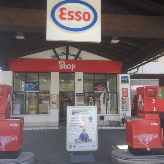 Eine Tankstelle mit einem ESSO-Schild über dem Laden. Vorne stehen zwei ESSO-Kraftstoffspender. Ein Schild steht zwischen ihnen.