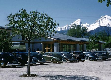 Ein Vintage-Bild von Oldtimern, die auf einem Parkplatz geparkt sind, mit einem verschneiten Berg im Hintergrund.