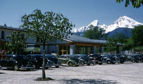 Ein Vintage-Bild von Oldtimern, die auf einem Parkplatz geparkt sind, mit einem verschneiten Berg im Hintergrund.