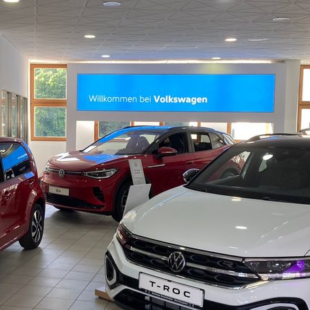 Ein Volkswagen-Autohaus präsentiert ein rotes und ein weißes Auto, mit einem blauen Schild an der Wand, das 'Willkommen bei Volkswagen' steht.