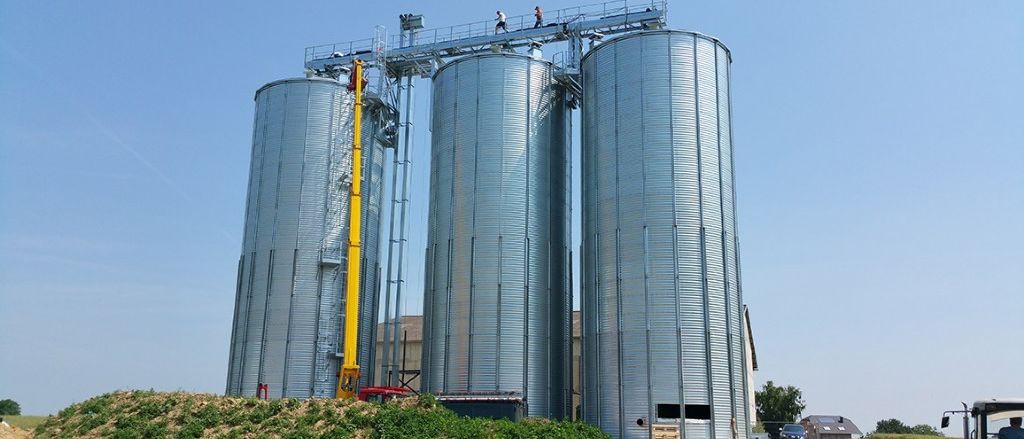 Drei Metallsilos auf einem Bauernhof. Ein gelber Kran steht links. Zwei Personen gehen auf dem Dach des Silos. Ein Haus steht rechts.