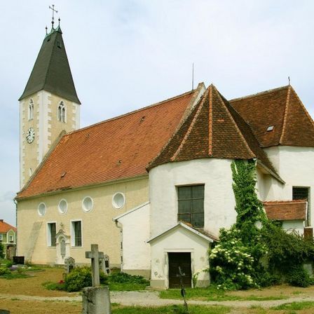 Eine alte Kirche mit einem braunen Dach und einem Turm mit einer Uhr. Das Gebäude ist von einem Friedhof mit einem Kreuz und Pflanzen umgeben.