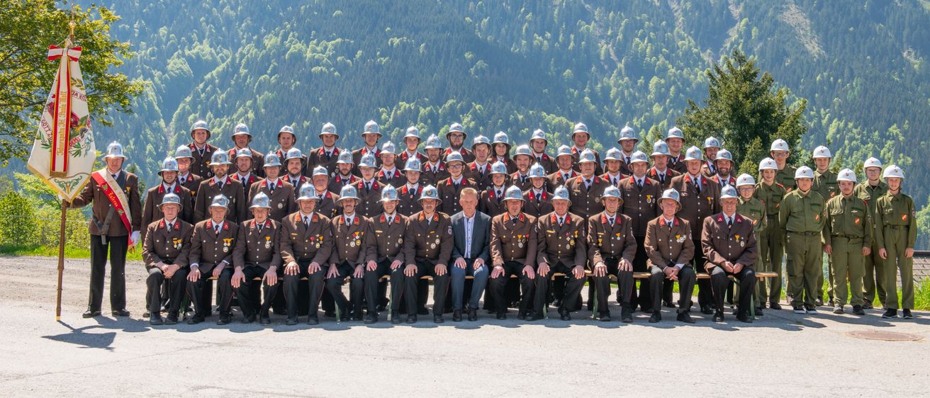 Eine Gruppe von Militärangehörigen in Uniform, mit Helmen, die in Reihen mit einem Gebirgshintergrund stehen und sitzen.