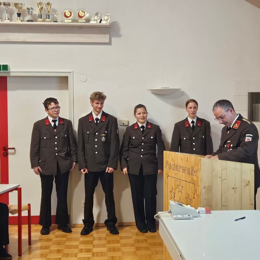 Five individuals dressed in uniforms stand by a wooden podium, likely attending a ceremony. Behind them, a wall holds a shelf with trophies and awards.