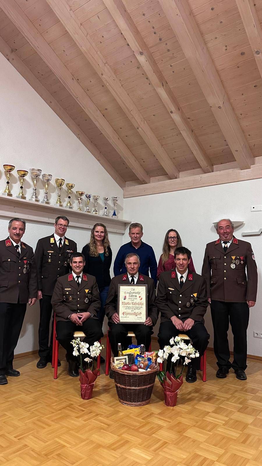A group of military personnel are posing for a photo with a framed certificate and flowers. They are wearing uniforms with medals. Behind them are trophies on a shelf.