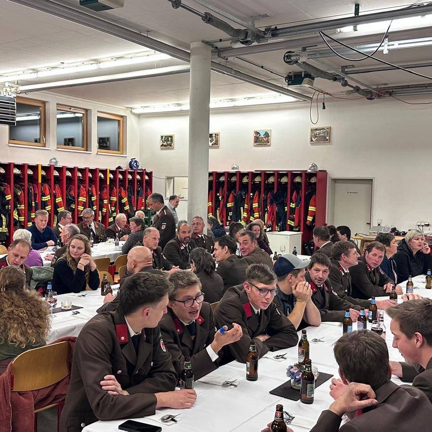 A large group of people, likely firefighters, are gathered indoors for a meeting or event. They sit at long tables, some with beer bottles, while others stand around. The room has mounted fire helmets and picture frames on the wall.