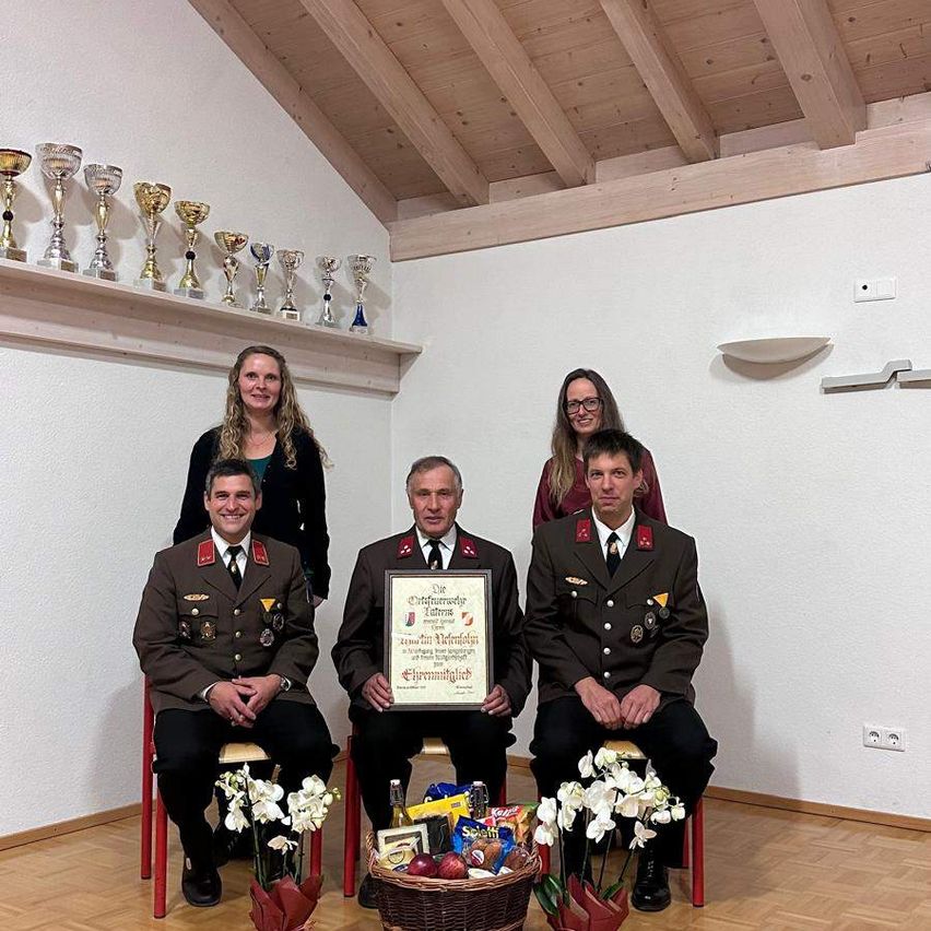 A group of people in military uniforms are posing for a photo. They are sitting on chairs in front of a basket filled with various items. Behind them, there are trophies on the wall and a white wall with a ceiling.
