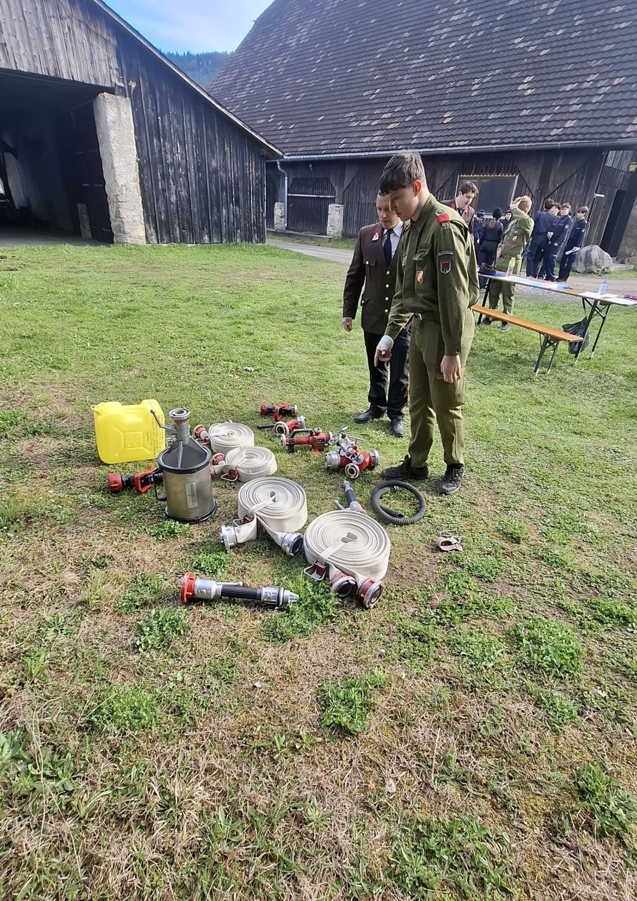Two men in uniforms standing on grass with fire extinguishers and hoses. A yellow container is nearby. Behind them, a group of people stands by a table with benches.