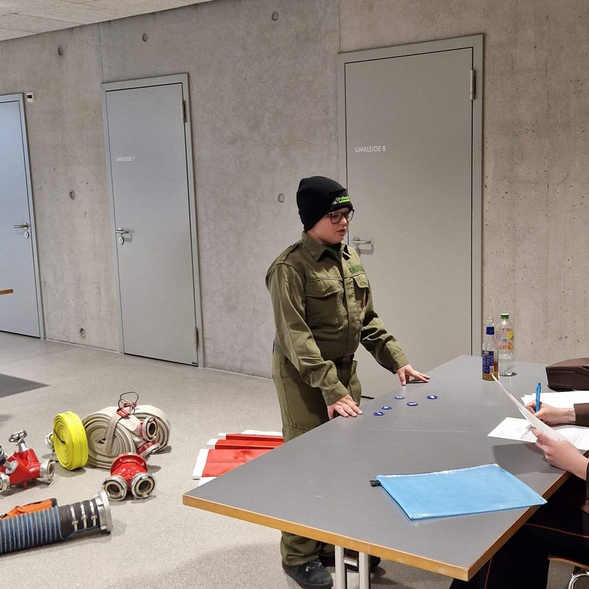 A person in uniform stands at a table, possibly for a drill. Doors are behind him, and firefighting equipment is on the floor. Another person sits at the table with papers.