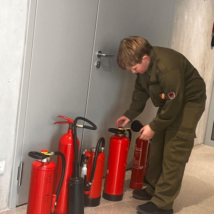 A man in a green uniform is inspecting a set of red fire extinguishers arranged against a gray door. He is holding one extinguisher and appears to be checking it.