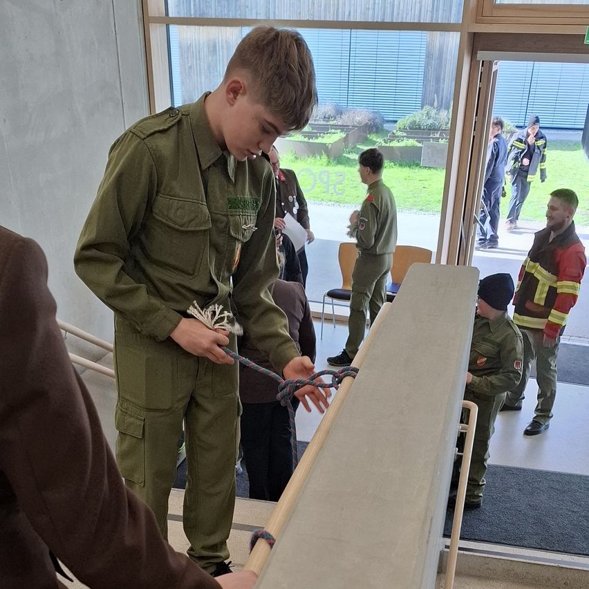 A young boy in a green uniform is demonstrating how to tie a knot while a group of people in uniform look on.