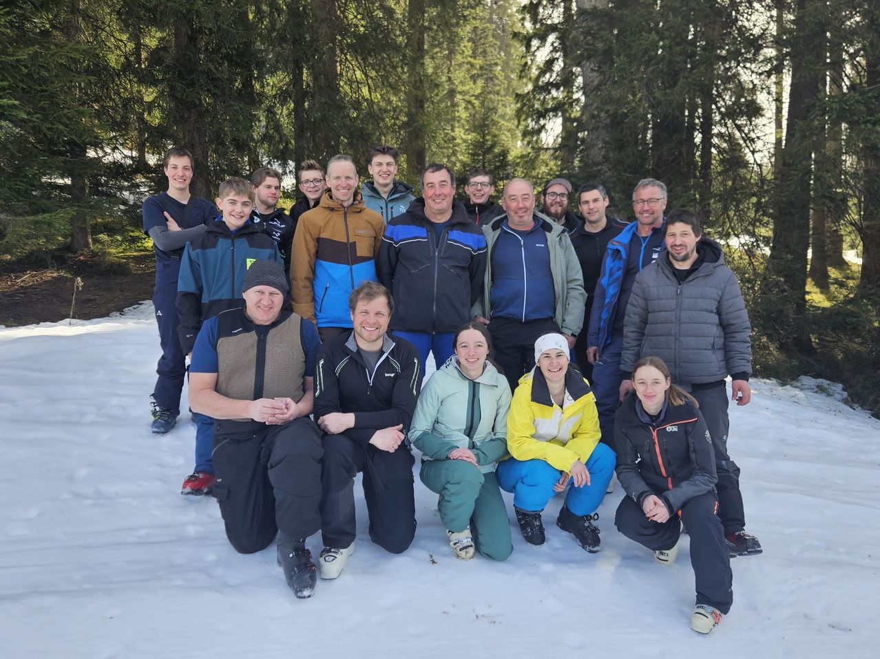Eine Gruppe von Menschen, die in Winterkleidung gekleidet sind und für ein Foto auf einer verschneiten Oberfläche mit hohen Bäumen im Hintergrund posieren.