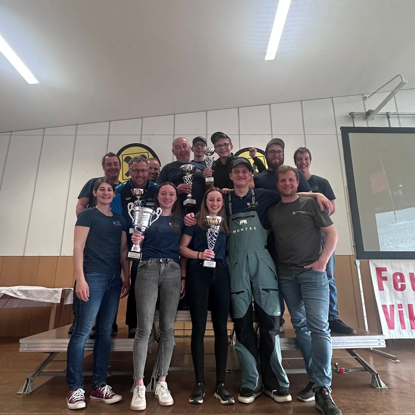 Eine Gruppe von Menschen, sowohl Männer als auch Frauen, posiert für ein Foto und hält Trophäen in den Händen. Sie alle lächeln und scheinen einen Sieg zu feiern.