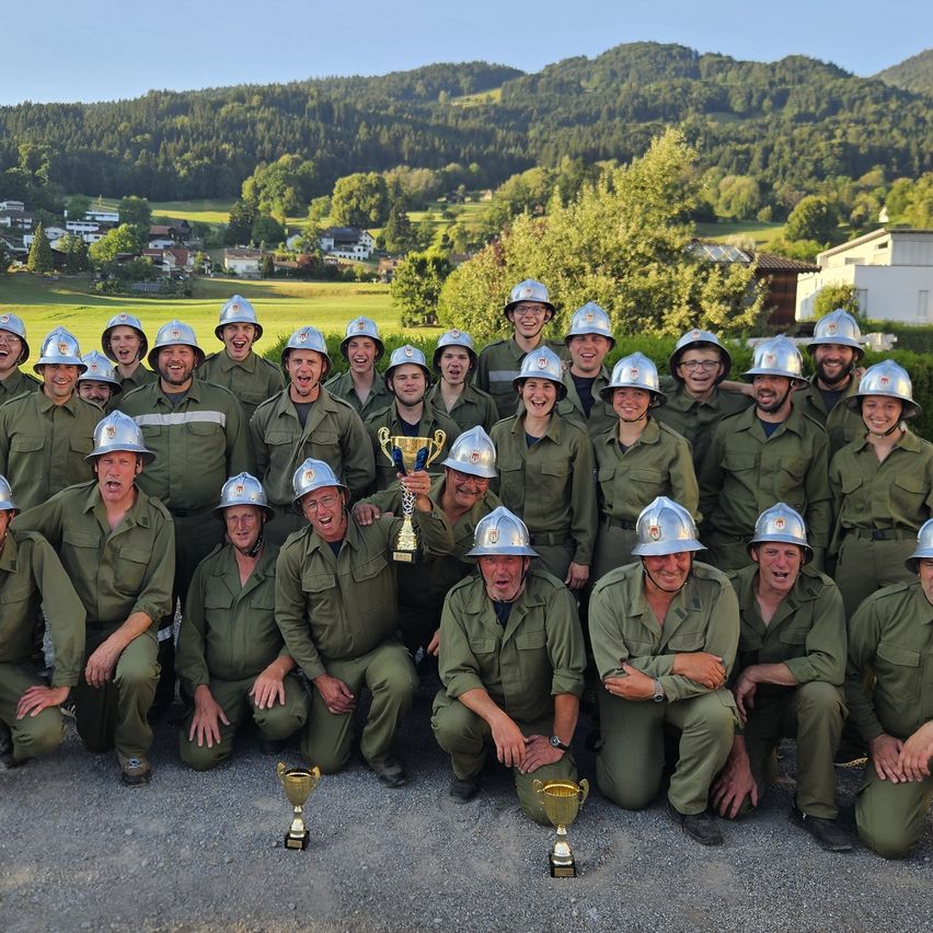Eine Gruppe von Menschen in grünen Uniformen und Helmen posiert für ein Foto mit einer Trophäe in der Mitte. Sie stehen und knien auf Kies mit einer malerischen Kulisse aus Bergen und Häusern.