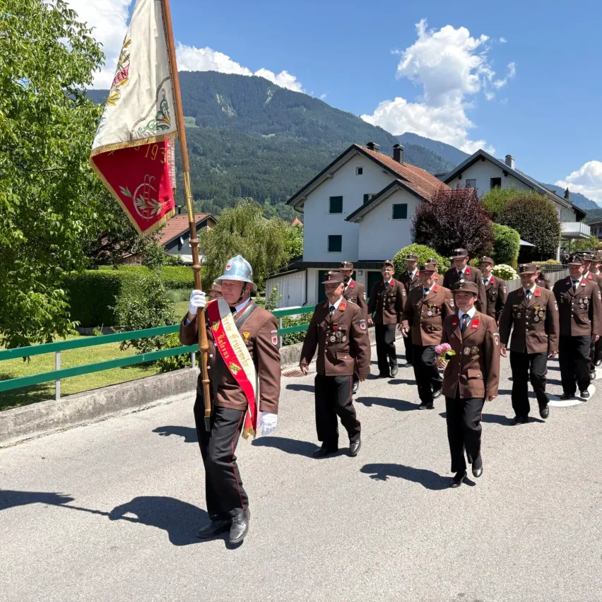 Eine Gruppe uniformierter Personen, die bei einer Parade marschiert, eine Fahne trägt und Mützen und Schuhe trägt. Sie gehen auf einer Straße mit Häusern und Grünflächen an den Seiten.