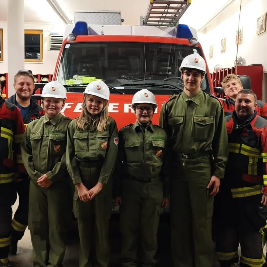 Eine Gruppe junger Menschen in grünen Uniformen und weißen Helmen steht vor einem roten Feuerwehrauto in einer Feuerwache.
