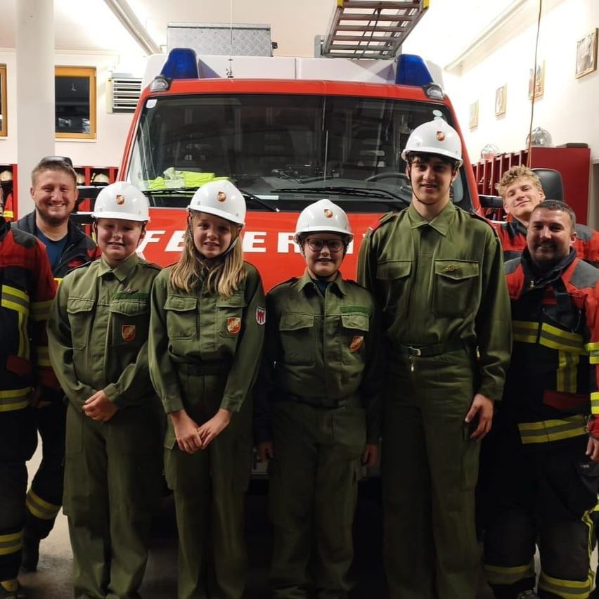 Eine Gruppe junger Menschen in grünen Uniformen und weißen Helmen steht vor einem roten Feuerwehrauto in einer Feuerwache.