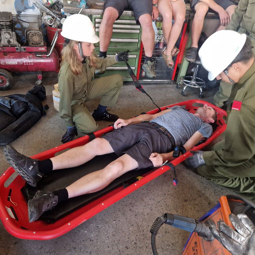 Ein Mann in einem roten Krankentrage wird von zwei Personen in grünen Uniformen und weißen Helmen betreut. Sie befinden sich in einer Werkstatt mit Werkzeugen und Ausrüstung im Hintergrund.