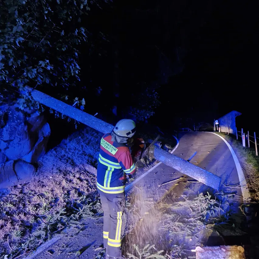 Ein Feuerwehrmann in voller Ausrüstung schneidet einen umgestürzten Ast mit einer Kettensäge in der dunklen Nacht.